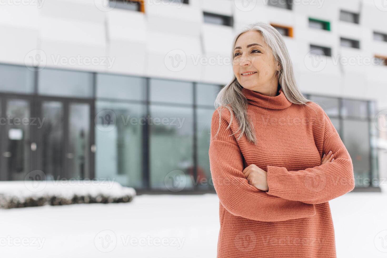 Senior woman with gray hair in a knitted sweater posing on the background of a modern house in winter photo