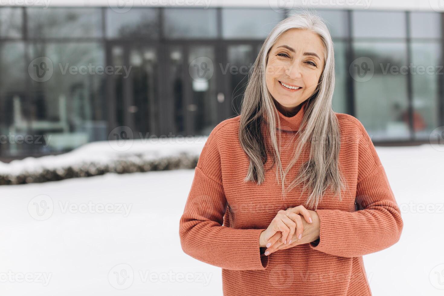 Senior woman with gray hair in a knitted sweater posing on the background of a modern house in winter photo