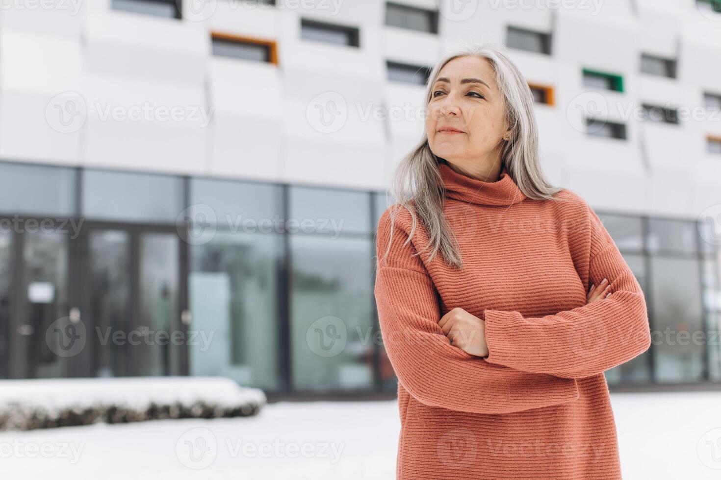 Senior woman with gray hair in a knitted sweater posing on the background of a modern house in winter photo