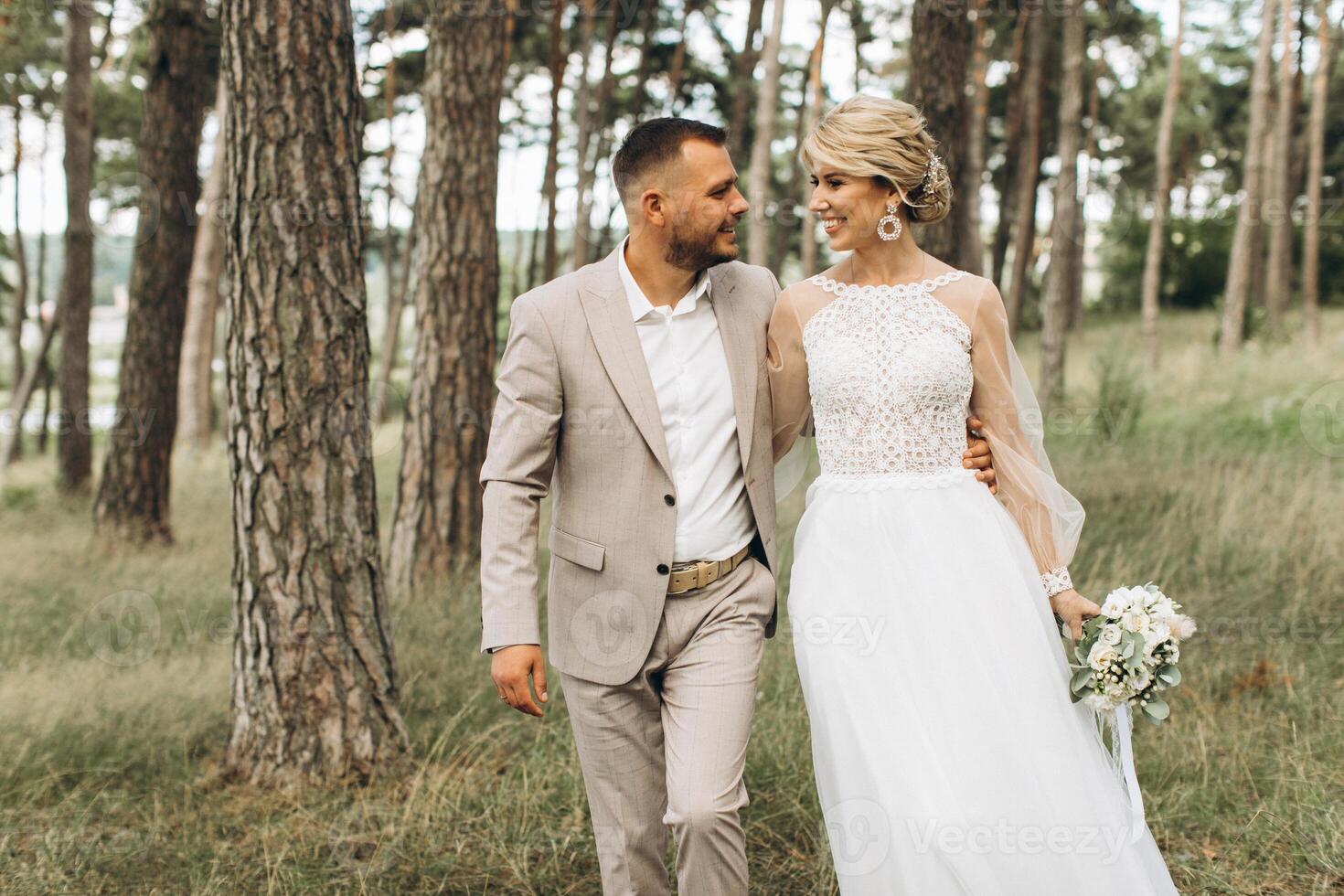 Wedding couple bride and groom walk hand in hand through a pine forest, sunlight filtering through the trees, highlighting their joyful expressions. photo