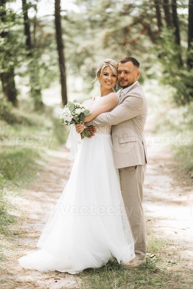 Wedding couple bride and groom hugging in pine forest, sunlight filtering through the trees, highlighting their joyful expressions. photo