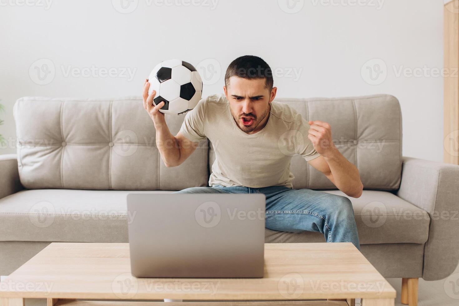 A man sits on a sofa in the living room holding a ball and cheers for the football team watching a match on a laptop photo