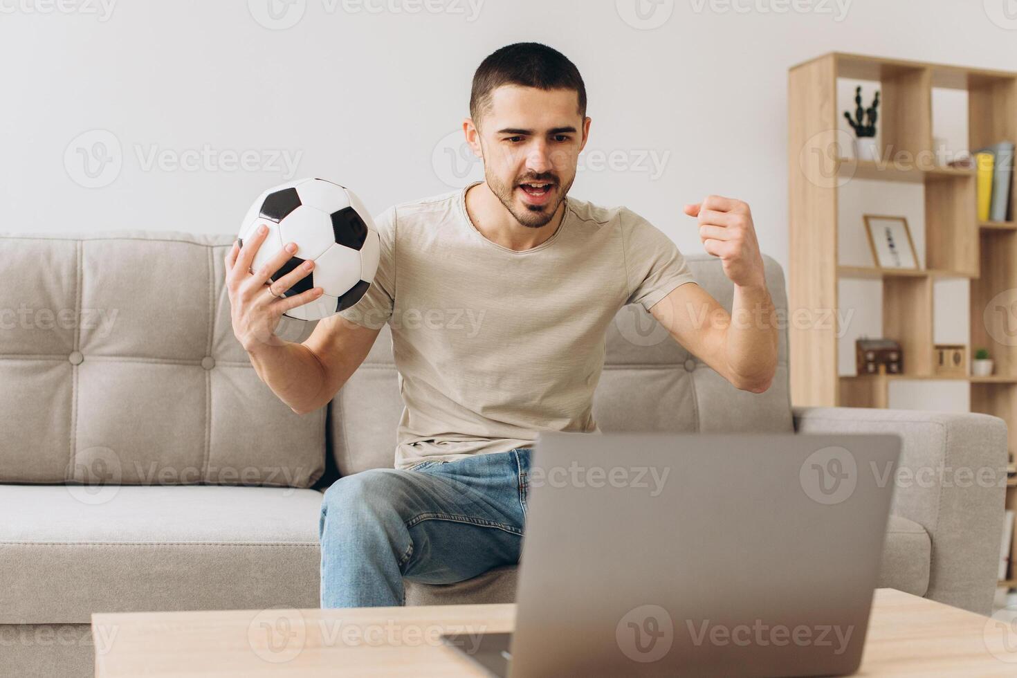 A man sits on a sofa in the living room holding a ball and cheers for the football team watching a match on a laptop photo