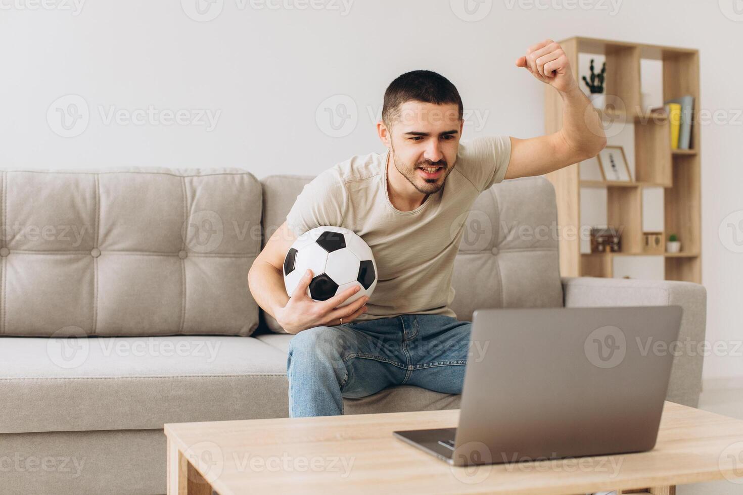 A man sits on a sofa in the living room holding a ball and cheers for the football team watching a match on a laptop photo