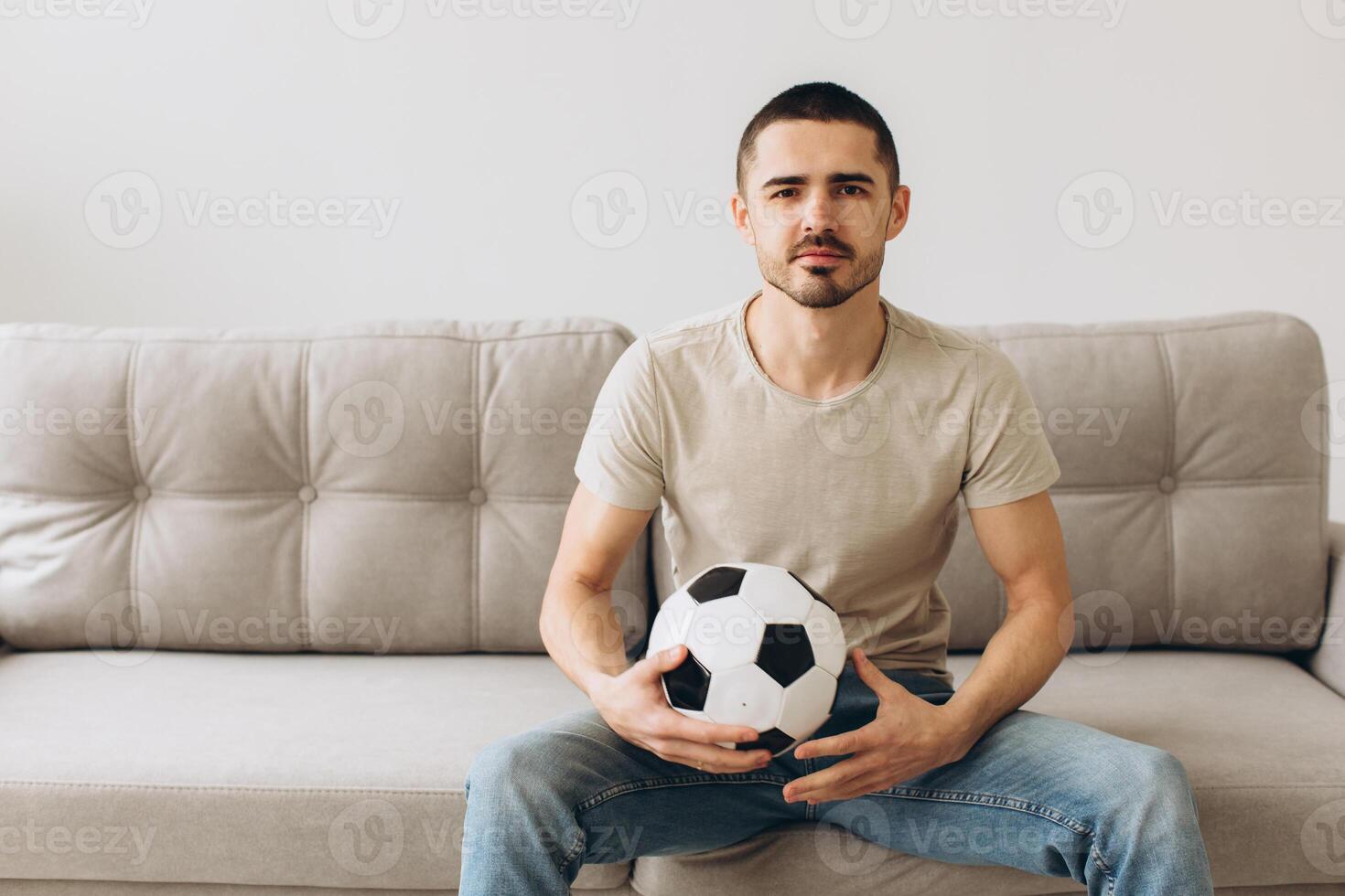 A man sits on a sofa in the living room holding a ball and cheers for the football team and watching a match photo