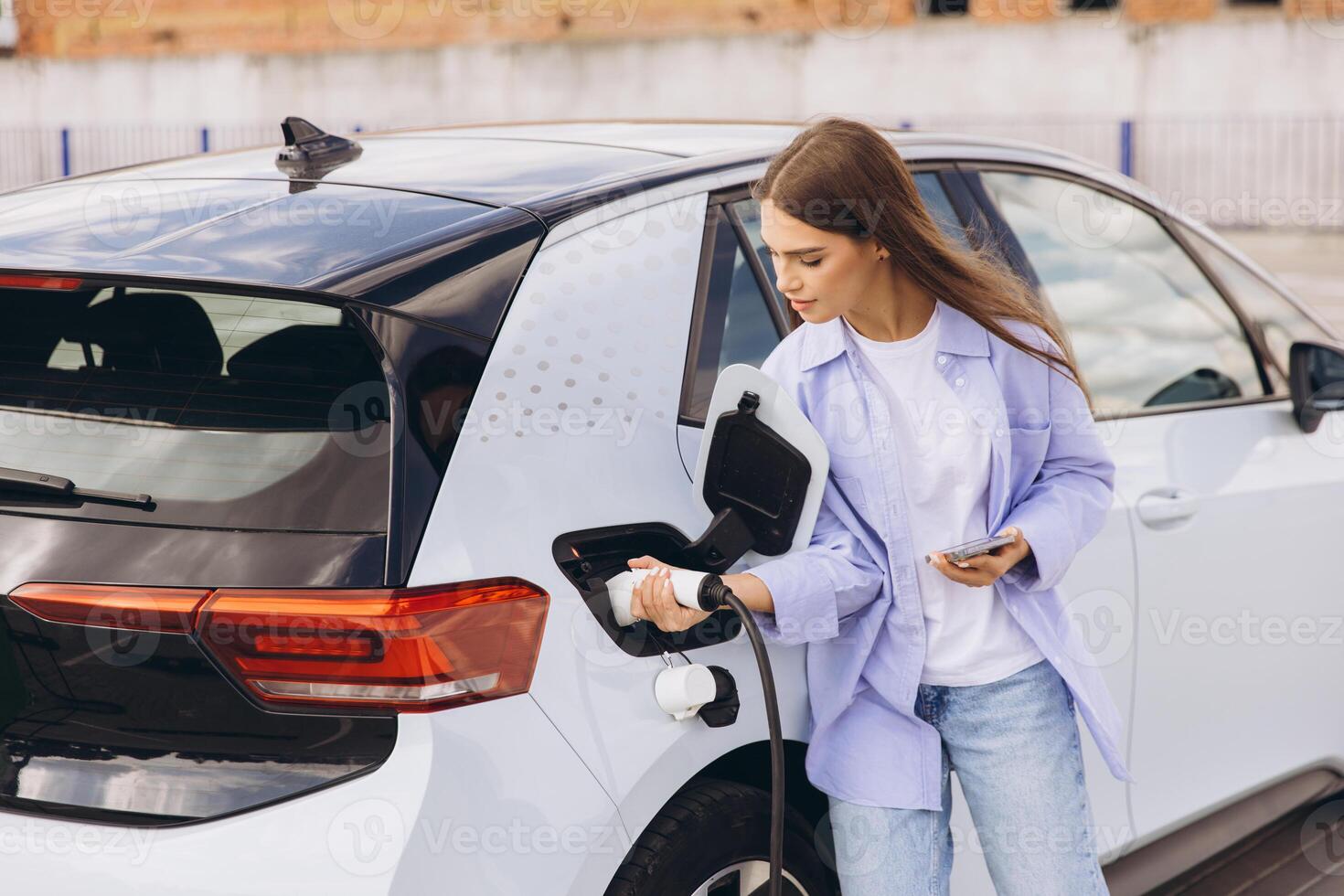 Young Woman Smiling While Charging Electric Car at Outdoor Charging Station on a Sunny Day photo