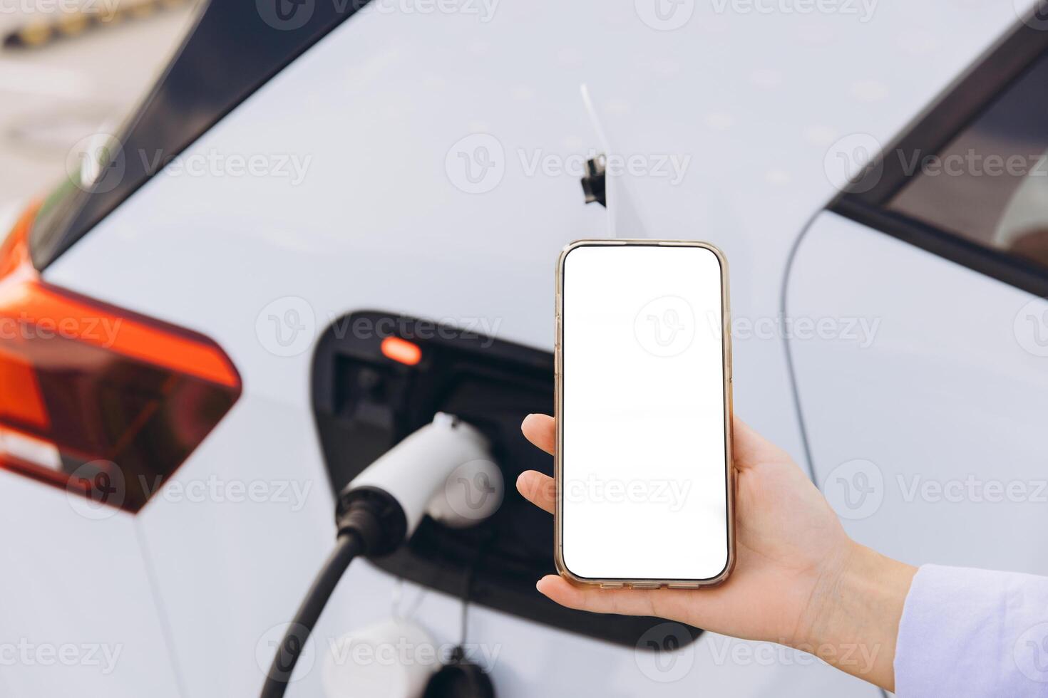 Smartphone in Hand Near Electric Car Charging at Station with Blank Screen for Mockup photo
