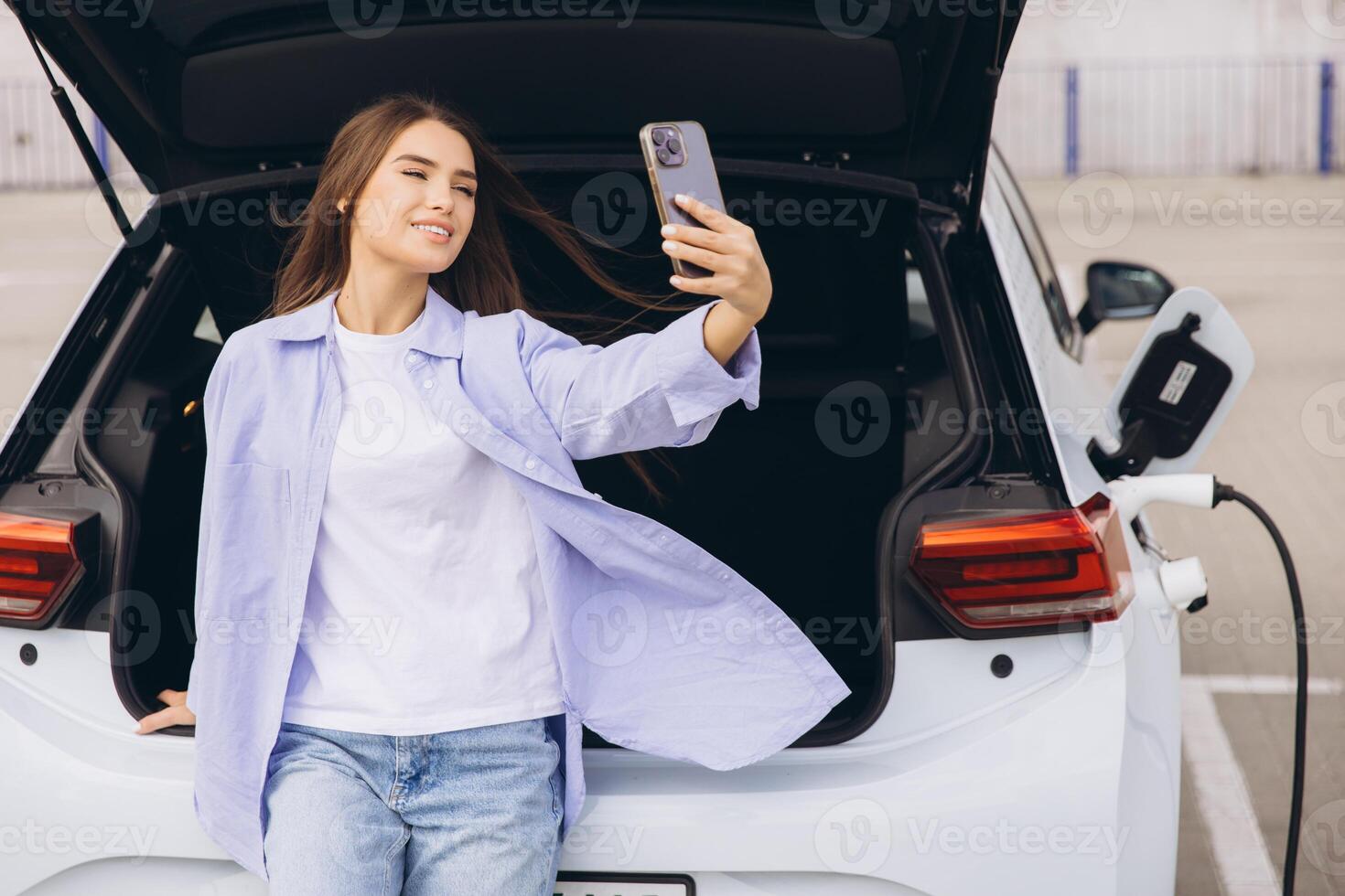 Young Woman Takes a Selfie Next to an Electric Car While Charging at a Modern Parking Lot photo