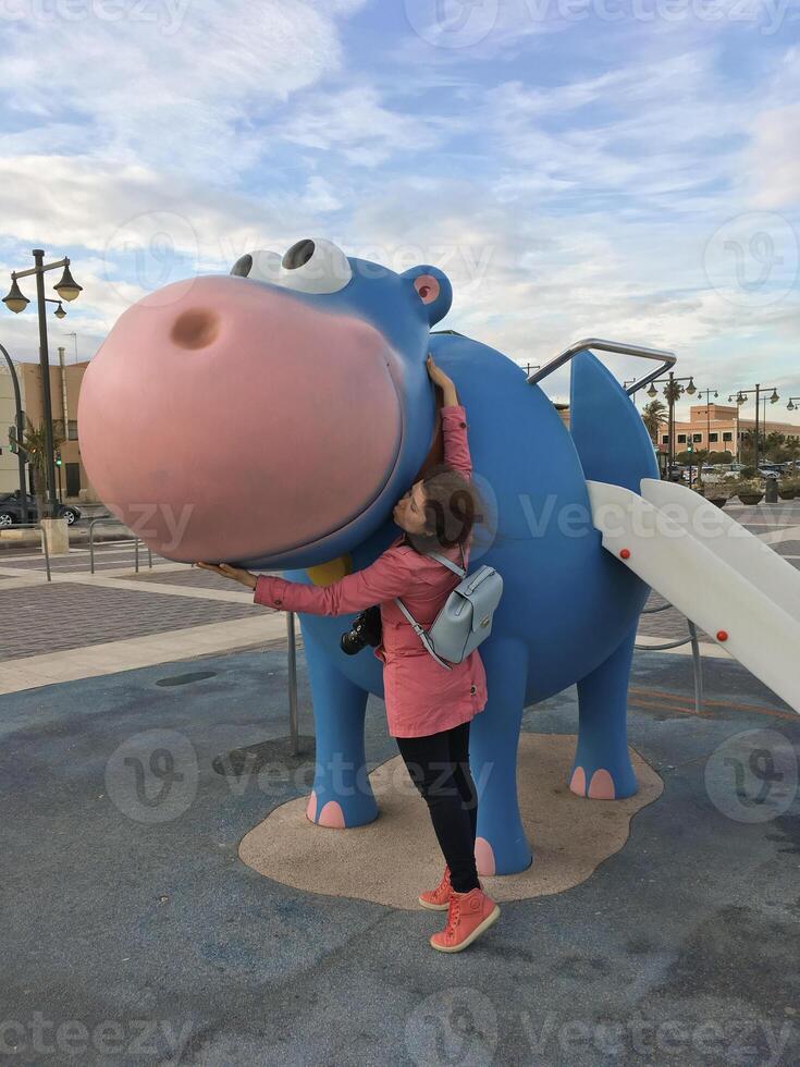 Young woman enjoying a delightful playful moment with a charming blue and pink hippo sculpture found in the playground photo