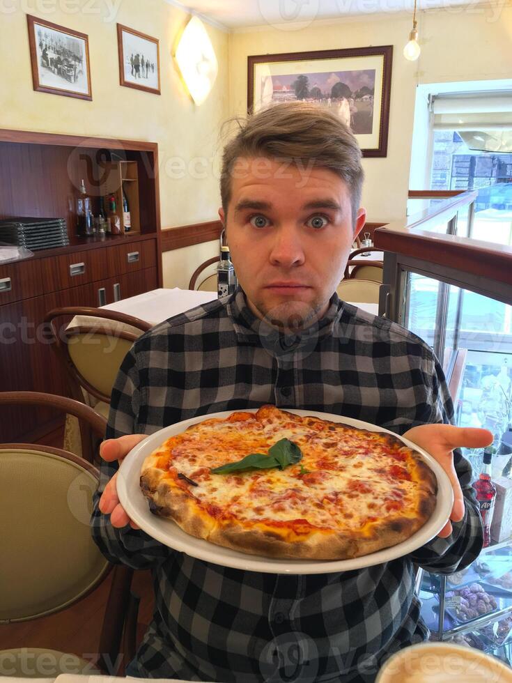 A young funny man with an astonished expression is holding a freshly baked pizza on a white plate in an indoor restaurant setting. Culinary Experience and Authenticity concept photo