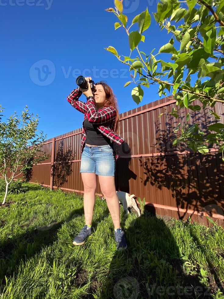 Millennial woman taking photo in a sunny backyard with a small tree and pet dog. Bright and clear blue sky providing a perfect backdrop for outdoor photography. Photographer, connection with nature