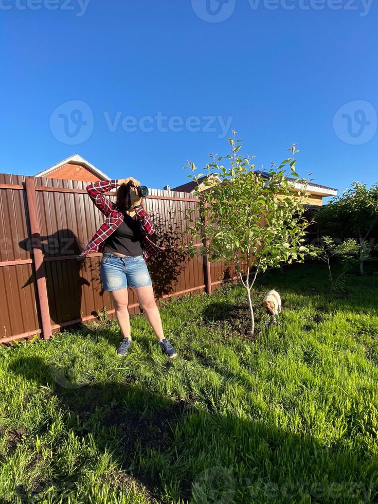 Millennial woman taking photo in a sunny backyard with a small tree and pet dog. Bright and clear blue sky providing a perfect backdrop for outdoor photography. Photographer, connection with nature
