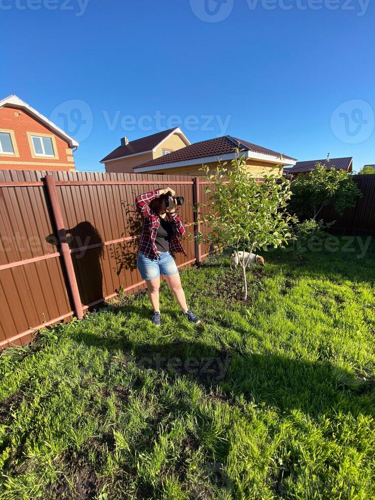 Millennial woman taking photo in a sunny backyard with a small tree and pet dog. Bright and clear blue sky providing a perfect backdrop for outdoor photography. Photographer, connection with nature