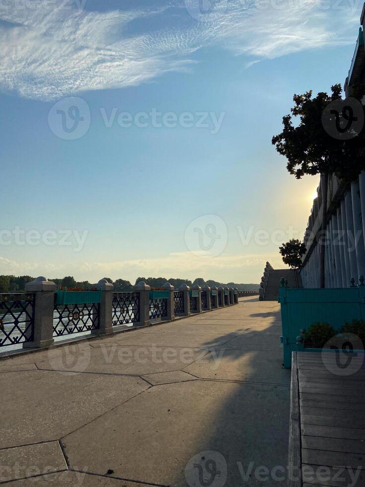 Scenic view of a riverside promenade with blue sky and fluffy clouds. Place leisurely walks and enjoying beautiful weather. Summer time and enjoying nature in urban settings photo