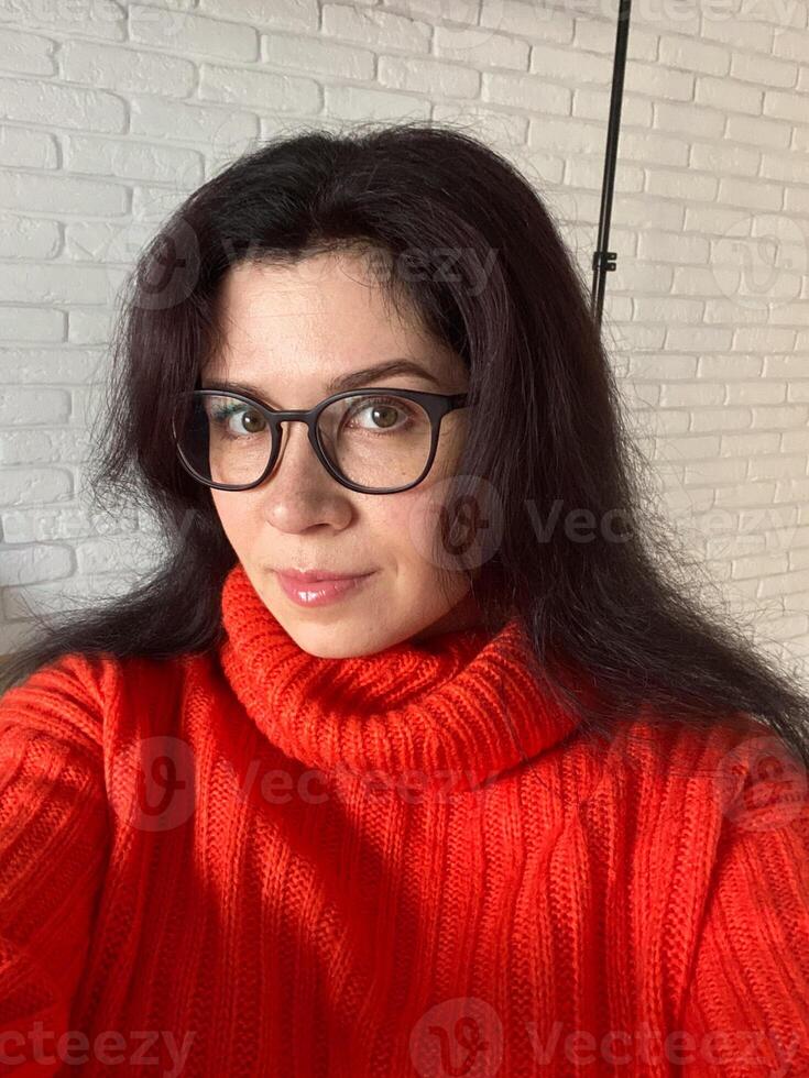 Selfie portrait woman in a vibrant red sweater poses confidently against a white brick wall. Glasses and dark hair create a striking contrast that exudes personal style and confidence photo