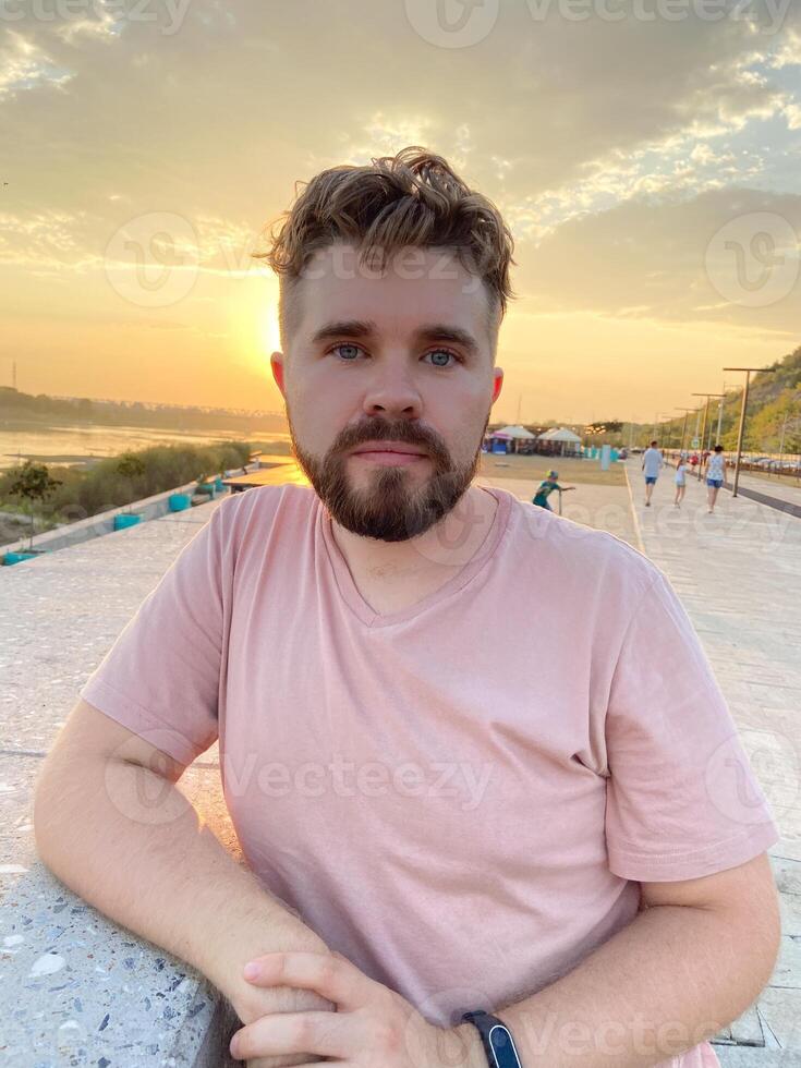 Bearded man gazes thoughtfully at the sunset, leaning against a concrete railing on a scenic promenade. Soft glow of the setting sun creates a serene and reflective atmosphere photo