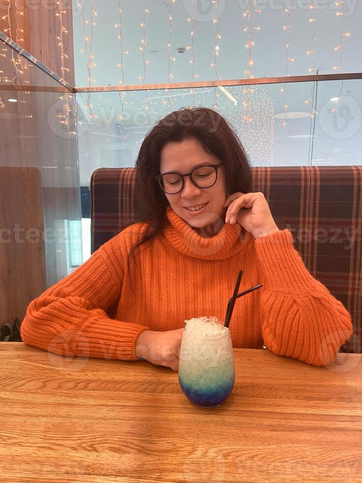Young woman in an orange sweater sits at a table, gazing happily at a colorful drink. Cozy atmosphere, with soft lighting and comfortable setting, enhances feeling of relaxation and enjoyment photo