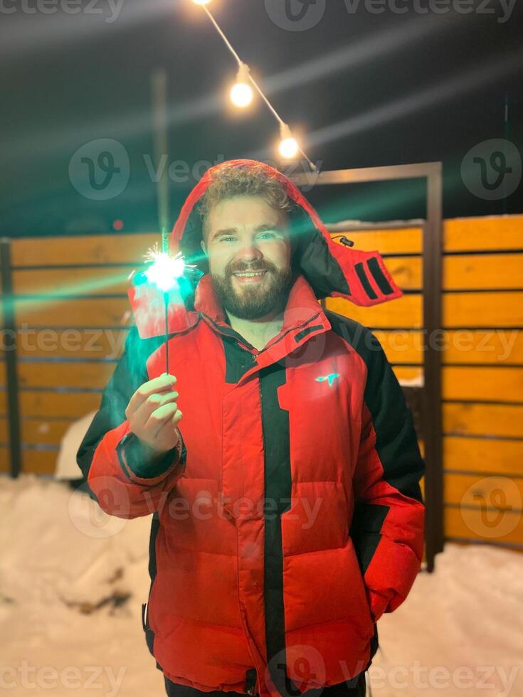 Bearded man in a red jacket holds a glowing sparkler, standing outside on a snowy winter night, with festive string lights hanging overhead. Christmas and New Year celebration concept photo