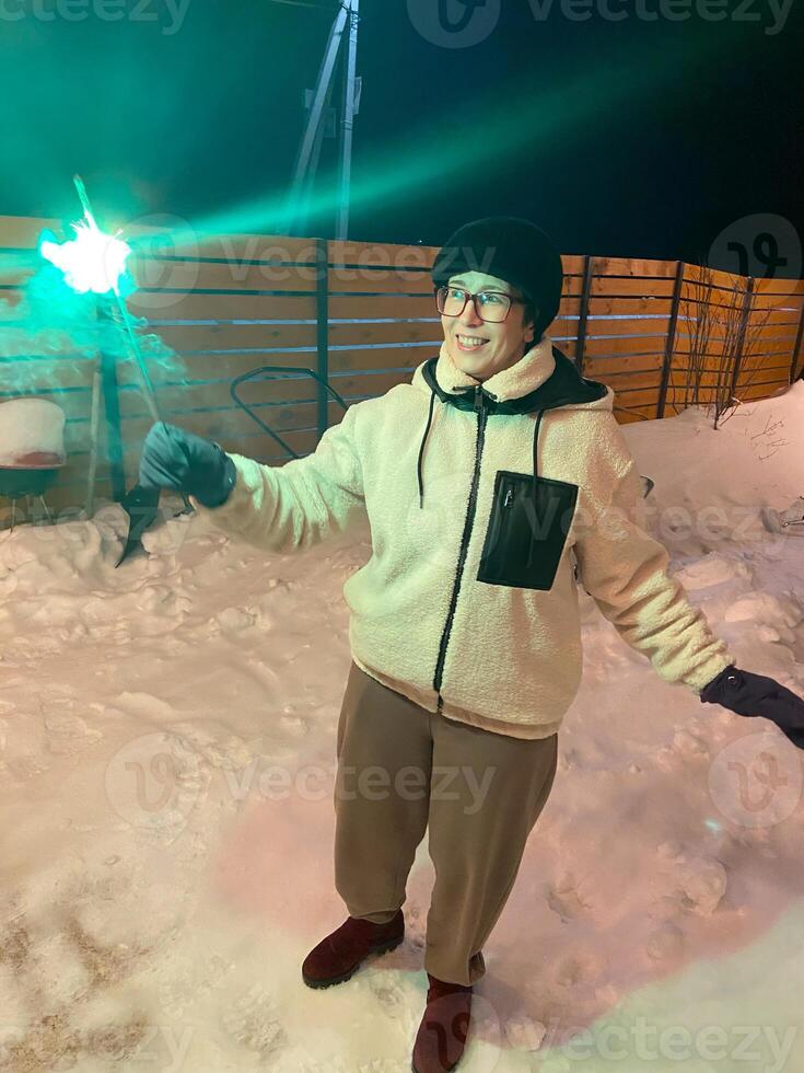 Woman in a red jacket holds a glowing sparkler, standing outside on a snowy winter night, with festive string lights hanging overhead. Christmas and New Year celebration concept photo