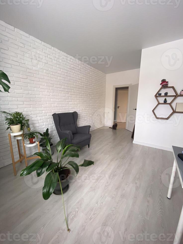 Stylish and minimalist living room features a comfortable armchair, vibrant green plants, and geometric wall shelves, creating a modern and serene atmosphere. The white brick wall photo