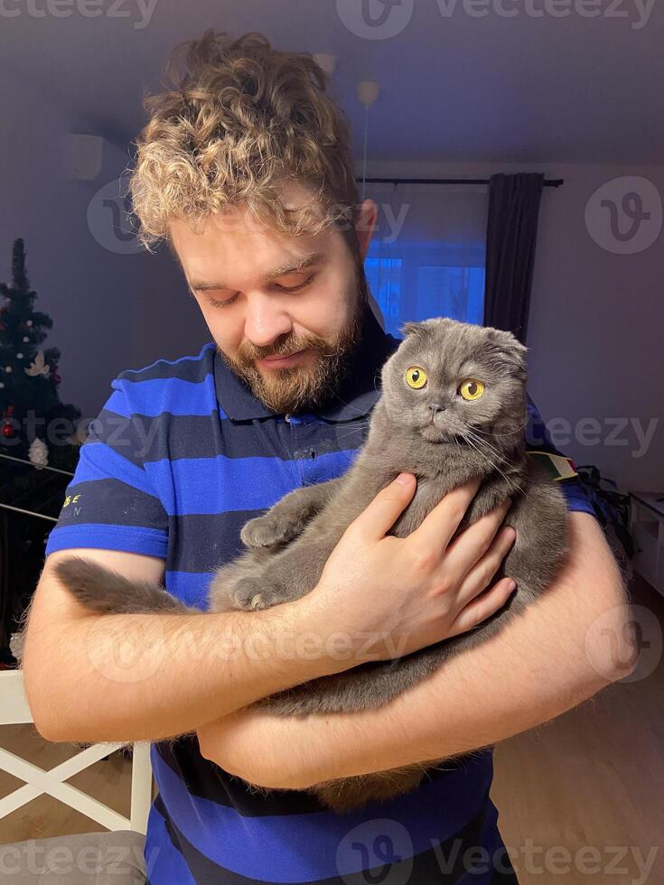 A millennial man is holding a Scottish Fold cat in his arms inside a warmly lit home decorated for Christmas holidays. Cat's striking yellow eyes making the moment intimate and endearing. photo