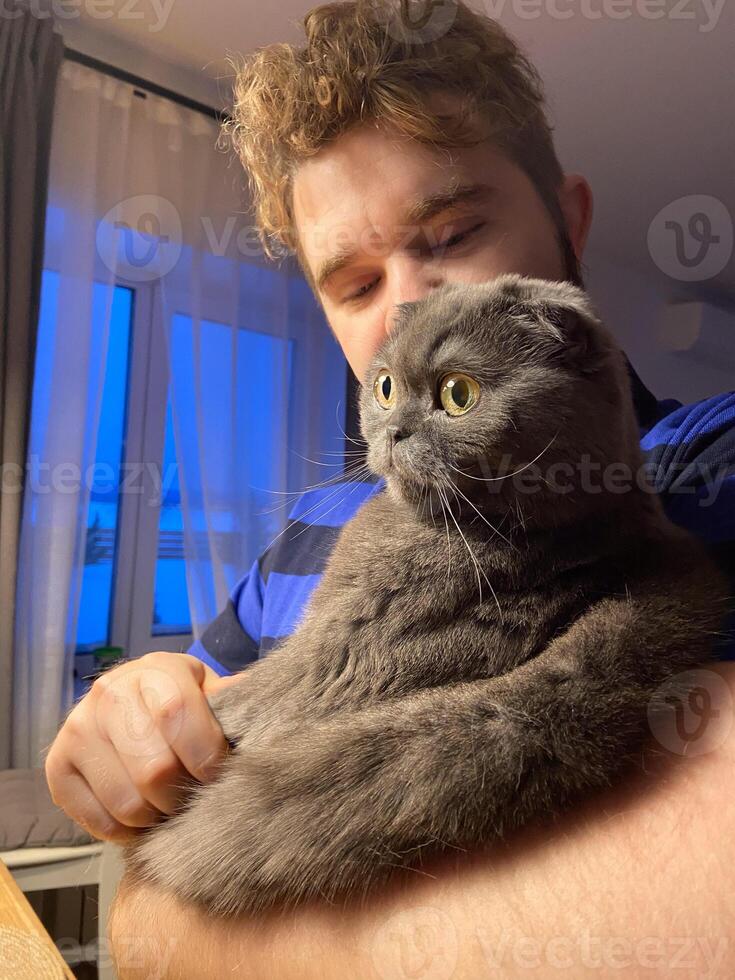 A millennial man is holding a Scottish Fold cat in his arms inside a warmly lit home decorated for Christmas holidays. Cat's striking yellow eyes making the moment intimate and endearing. photo