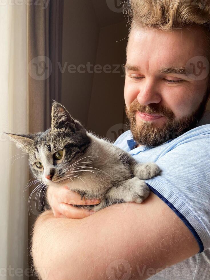 Millennial man gently holds a cat by a sunlit window, capturing a moment of serene companionship. Warm light, sense of peace and connection between human and pet. photo