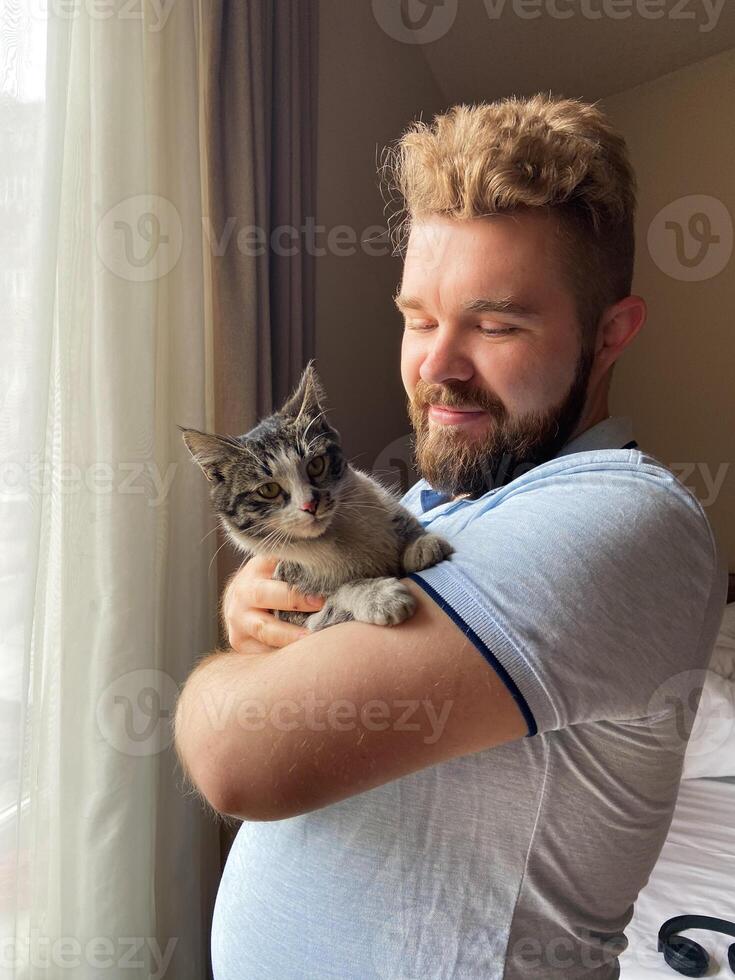Millennial man gently holds a cat by a sunlit window, capturing a moment of serene companionship. Warm light, sense of peace and connection between human and pet. photo