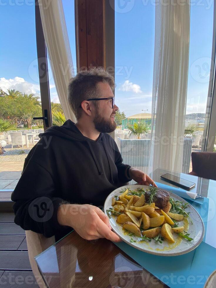 Bearded man enjoys a meal at a seaside restaurant, with a plate of steak and potatoes. Bright and open atmosphere, surrounded by palm trees and a scenic view of the beach outside photo