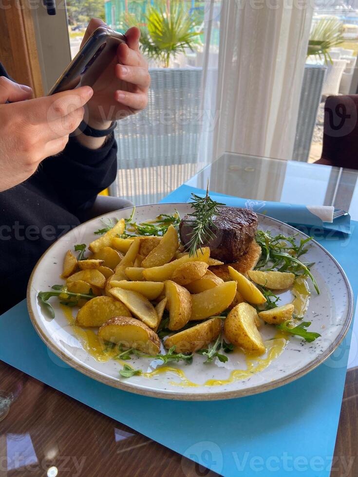 Close-up man takes a photo of a dish consisting of grilled meat and potatoes. Social networks, blog and food blogger concept