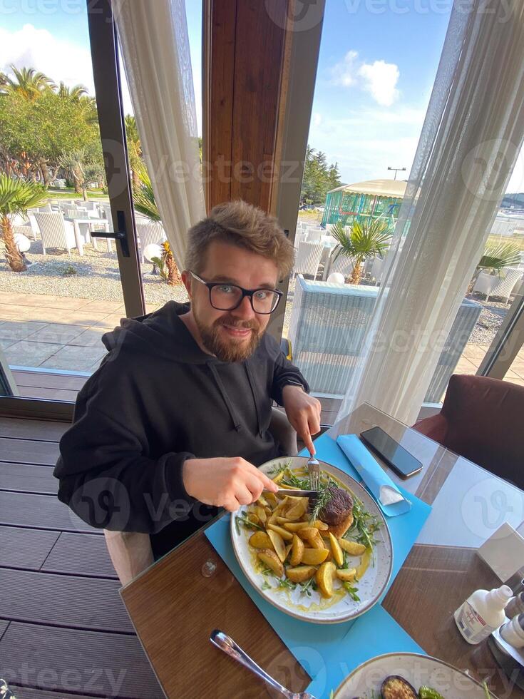 Bearded man enjoys a meal at a seaside restaurant, with a plate of steak and potatoes. Bright and open atmosphere, surrounded by palm trees and a scenic view of the beach outside photo