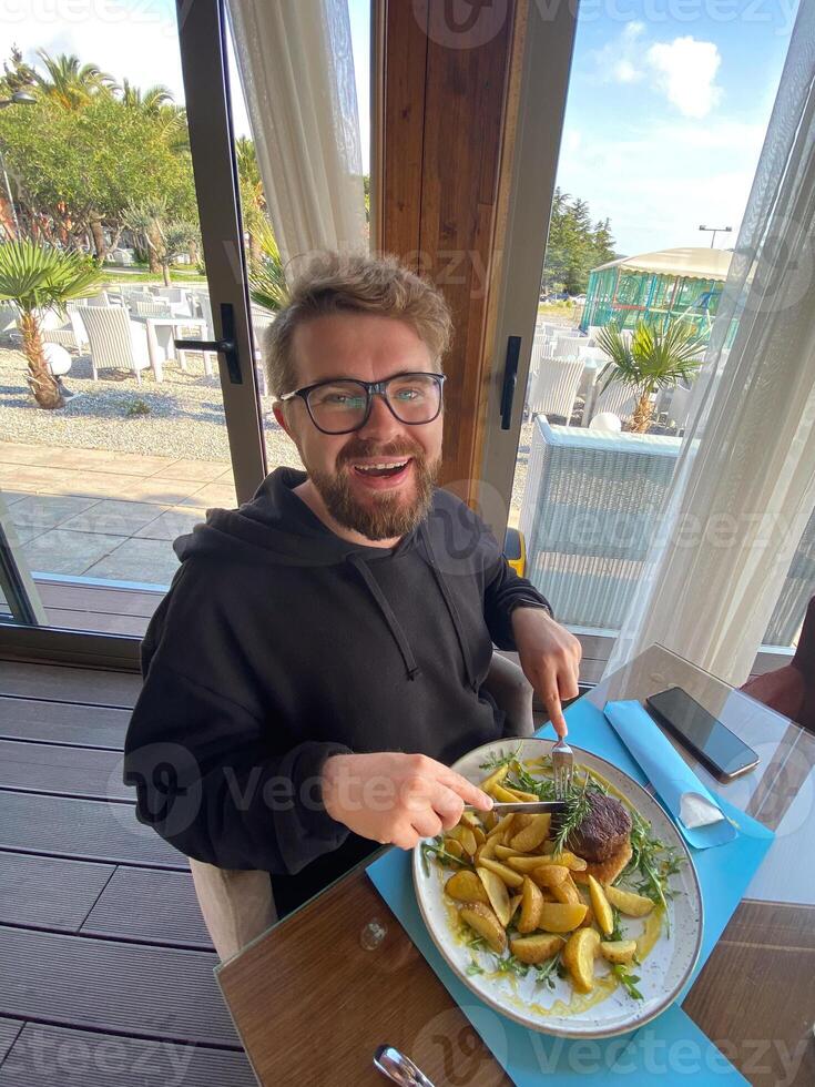 Bearded man enjoys a meal at a seaside restaurant, with a plate of steak and potatoes. Bright and open atmosphere, surrounded by palm trees and a scenic view of the beach outside photo