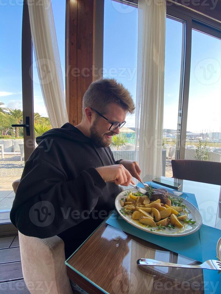 Bearded man enjoys a meal at a seaside restaurant, with a plate of steak and potatoes. Bright and open atmosphere, surrounded by palm trees and a scenic view of the beach outside photo