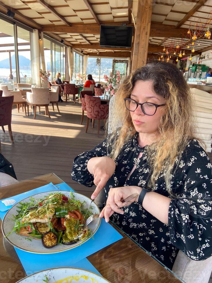 Woman enjoys a meal at a seaside restaurant, with a plate of grilled vegetables and chicken on toast. Bright and open atmosphere, surrounded by palm trees and a scenic view of beach outside photo