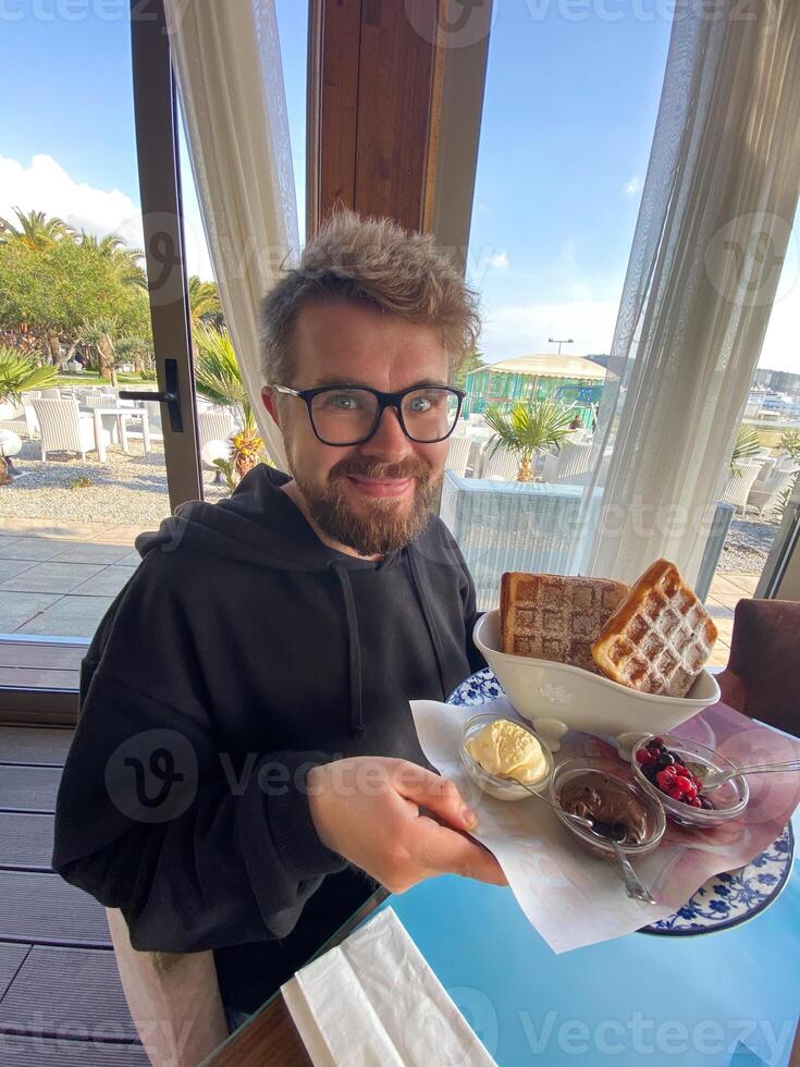 Man smiles while holding a plate of waffles with sides of ice cream, berries, and chocolate at a seaside restaurant. Bright and sunny setting, with palm trees visible outside, photo