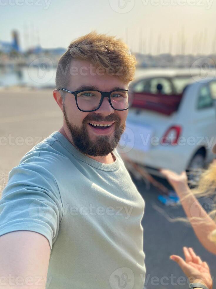 Joyful man takes a selfie by a car at the seaside. Male smiling broadly, enjoying a sunny day by the water. photo