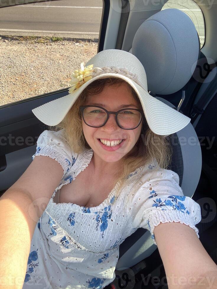 Cheerful woman in a white dress and wide-brimmed hat smiles warmly while sitting in a car, exuding joy and a sense of adventure. Bright sunlight accentuates her contentment photo
