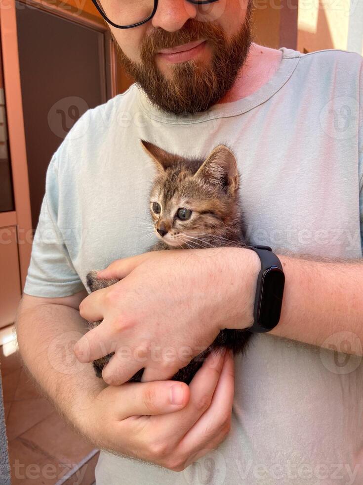 Millennial man in a straw hat and sunglasses holds lovingly at a small kitten in his hands. Tenderness moment captures the bond between human and pet. Themes of care and affection photo