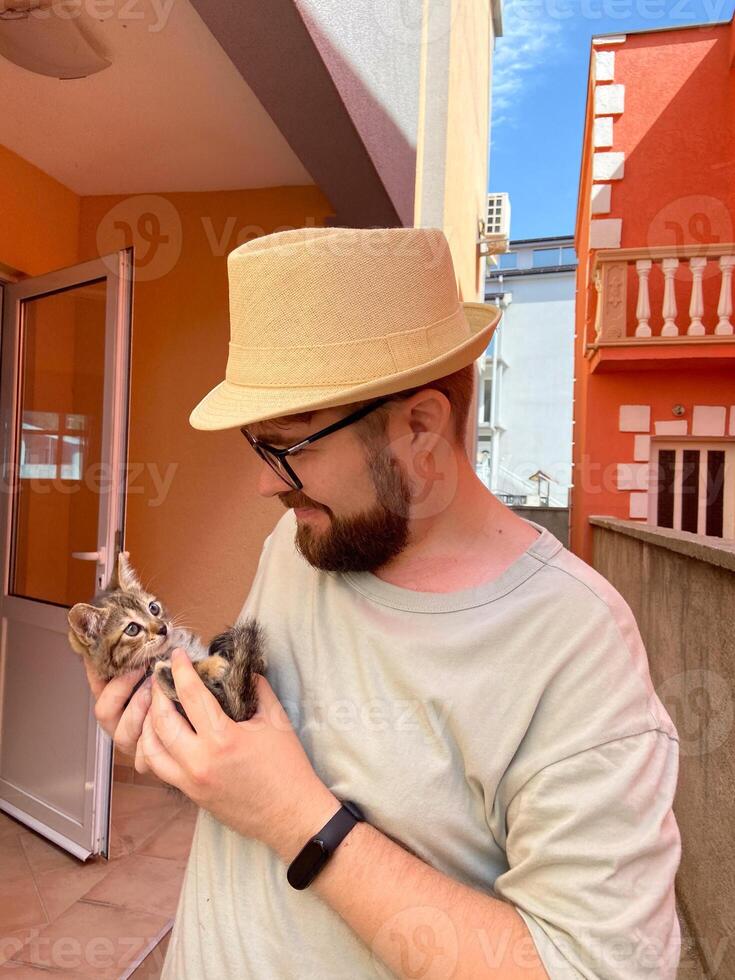 Millennial man in a straw hat and sunglasses holds lovingly at a small kitten in his hands. Tenderness moment captures the bond between human and pet. Themes of care and affection photo