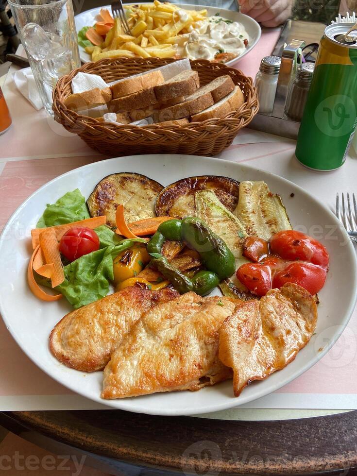 Delicious meal featuring grilled vegetables and chicken breast is elegantly presented on a white plate. Basket of sliced bread and a soft drink can, creating a cozy dining atmosphere photo