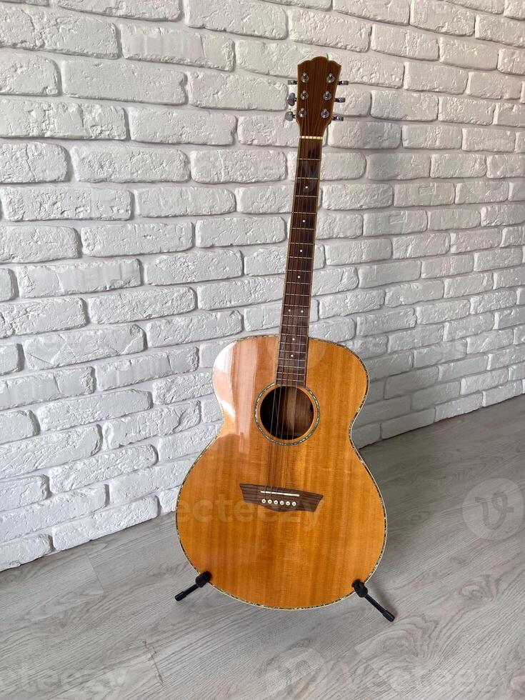 Wooden acoustic guitar stands elegantly against a textured white brick wall, creating a harmonious blend of music and art. Instrument craftsmanship photo