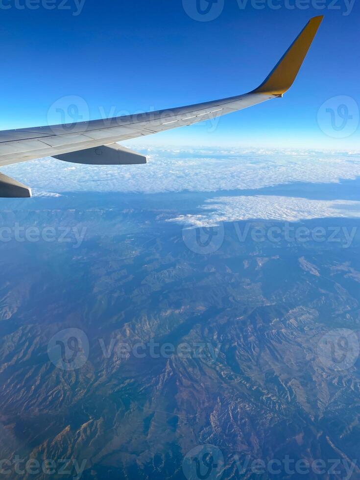 View of airplane wing through window at high altitude, showcasing clear blue sky and clouds far below. Serene beauty of flight and the expansive view of the horizon from above. photo