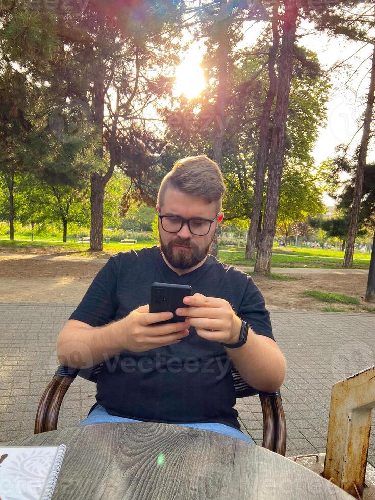 Bearded man sits in a park, focused intently on smartphone with sun shining through the trees in background. Peaceful setting highlights the blend of technology photo