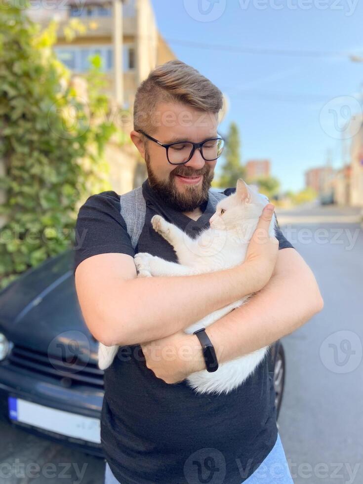 Bearded man gently embraces a white cat, conveying a moment of warmth and connection with pet. Outdoor setting adds an element of everyday life and companionship photo