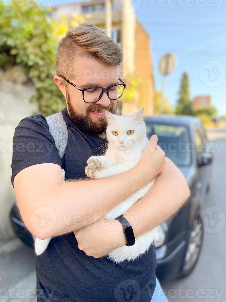 Bearded man gently embraces a white cat, conveying a moment of warmth and connection with pet. Outdoor setting adds an element of everyday life and companionship photo