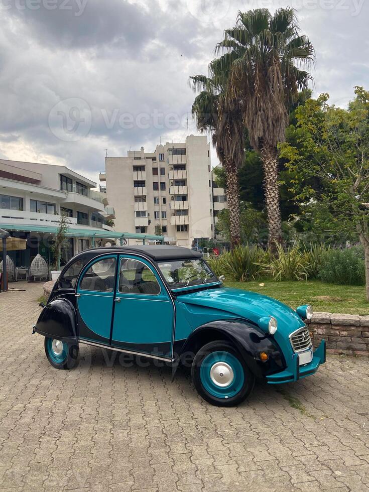 Classic vintage car rests in an urban setting, juxtaposing modern architecture with nostalgic charm. Bright teal vehicle stands out against backdrop of palm trees photo
