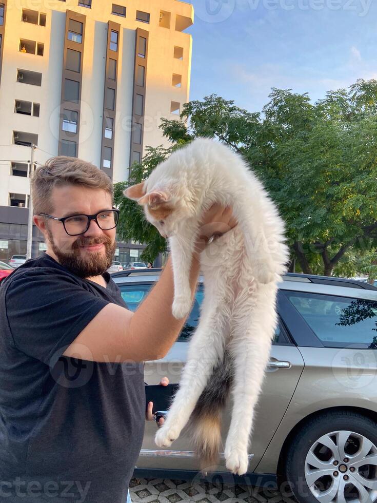 Bearded man joyfully lifts a fluffy white cat in an urban setting, with a high-rise building and leafy trees in background. Playful moment captures bond between pets and their owners photo
