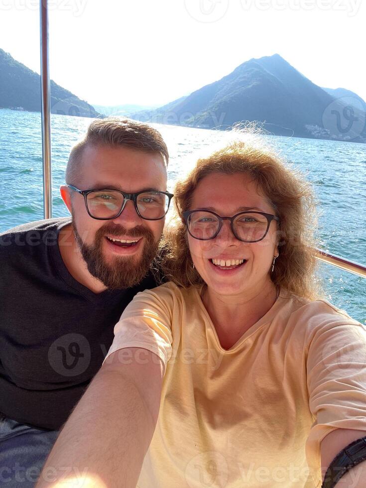 Happy couple taking selfie on smartphone and enjoys a scenic boat ride with stunning mountain views in background. Smiles reflect the joy and freedom of a beautiful adventure on the water. photo