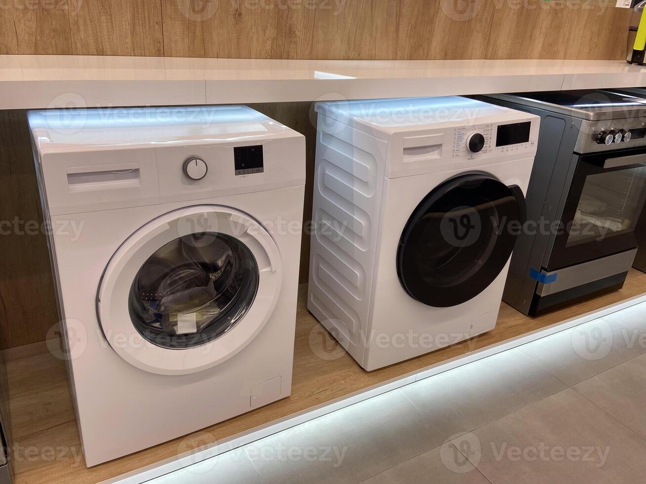 Row of modern washing machines is displayed in an electronics store, showcasing the latest models with sleek designs and advanced features. Under bright lights, the machines stand photo