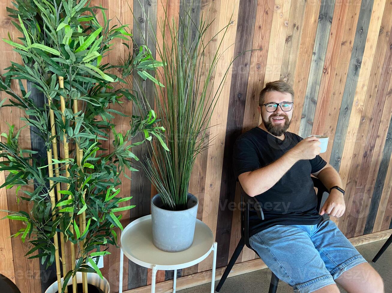 Bearded man in casual attire sits on a chair, holding a cup of coffee, against a stylish wooden wall with potted plants. Relaxed and modern atmosphere, perfect for enjoying a peaceful moment photo